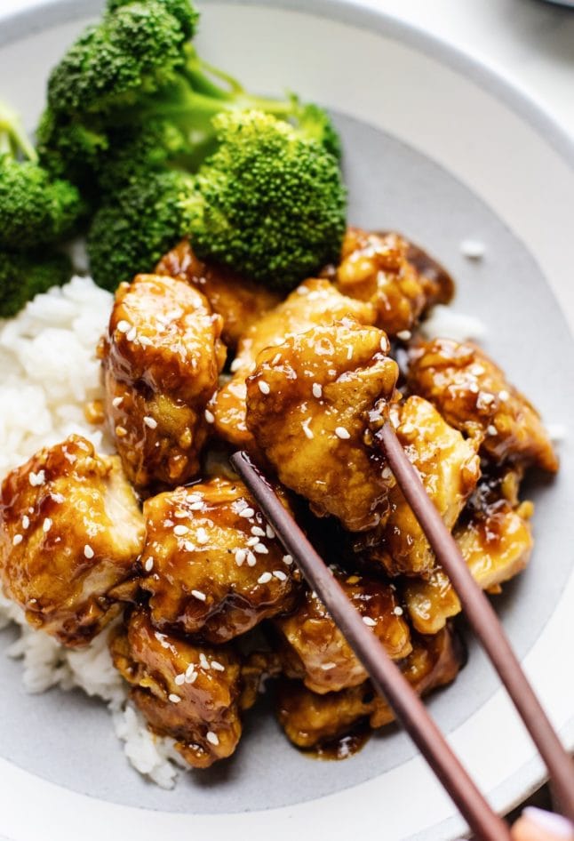 A close up of chopsticks picking up a piece of honey sesame chicken with broccoli and rice in the background.