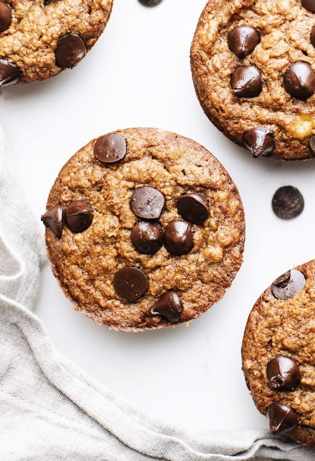 Vegan banana muffins with chocolate chips on top arranged on a white counter with a gray napkin in the corner.