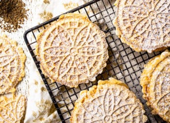 Overhead image of pizzelle on a wire cooling rack with whole anise seeds and powdered sugar.