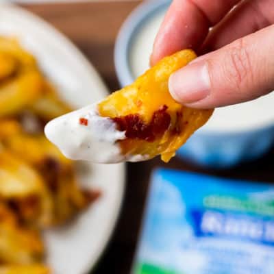 A french fry covered in cheese, bacon, and Ranch dressing being held up with a plate of fries, a packet of Ranch dressing, and a small bowl of Ranch in the background