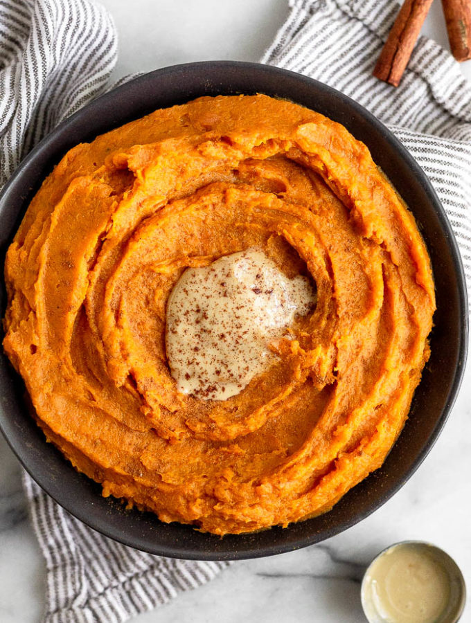 A bowl of tahini whipped sweet potatoes with a dollop of tahini on top and cinnamon sprinkled over them. The bowl is on a towel and next to it is a tablespoon with a little tahini in it and cinnamon sticks.