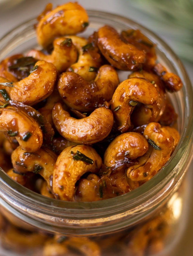 A jar filled with rosemary roasted cashews. Behind it is rosemary and a tan linen.