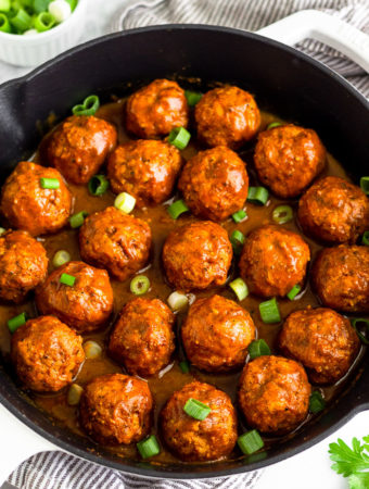 A large cast iron skillet filled with BBQ chicken meatballs covered in BBQ sauce and topped with green onions. Around it is some fresh parsley and a bowl of chopped green onions.