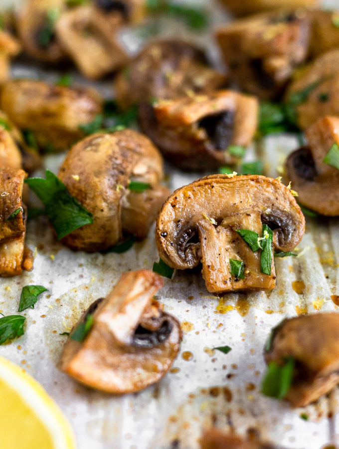Close up of a balsamic roasted mushrooms on a baking sheet surrounded by more mushrooms. They are topped with fresh parsley.