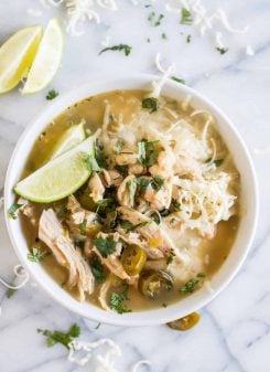 A bowl filled with white chicken chili, topped with cheese, cilantro and lime wedges.
