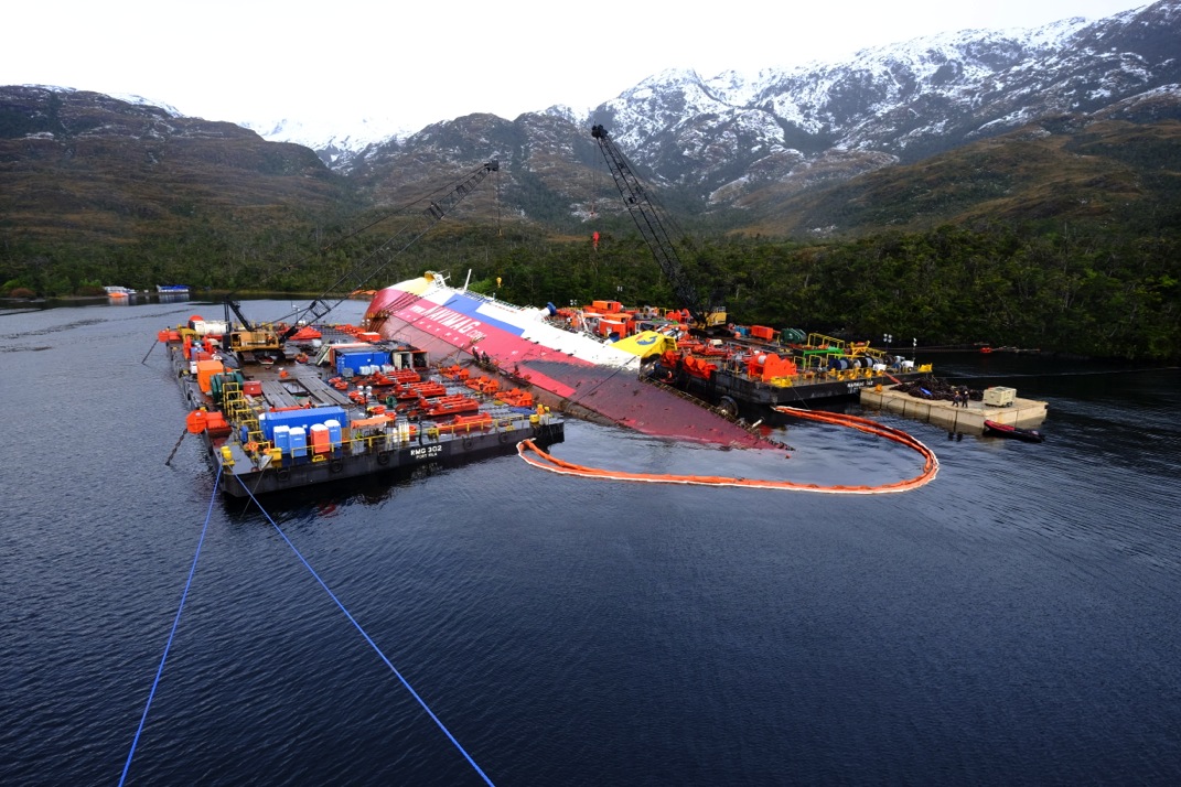 The Amadeo 1 lies sideways on her starboard side in water. She is attended to by two floating docks on either side