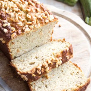 A keto zucchini bread topped with hazelnuts with two slices cut off on a wooden board and a knife
