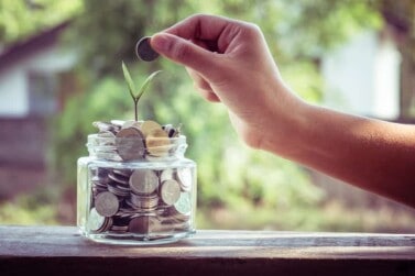 hand putting coins into glass jar with seedling growing out of it