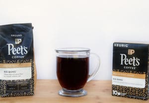 Bag of Peet's Coffee and box of Peet's Coffee K-Cups with a glass mug of black coffee sitting on a cutting board