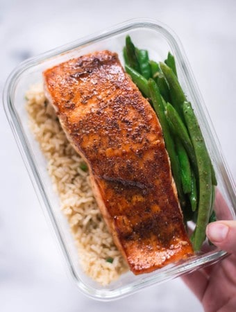 Hand holding a single meal prep container which has pan-roasted salmon, garlic rice, and parmesan garlic green beans for how to meal prep salmon.