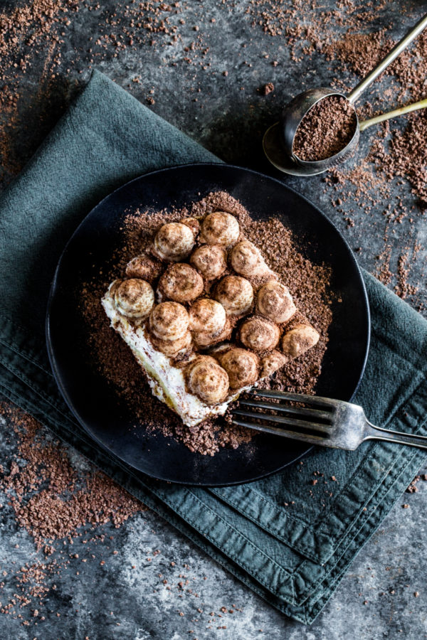 A square slice of tiramisu served on a black plate with an antique fork.