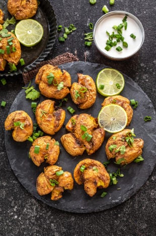 masala smashed potatoes served on a circular cheese board with lime wedges placed on the side. There's also a white color dip in a bowl in the background