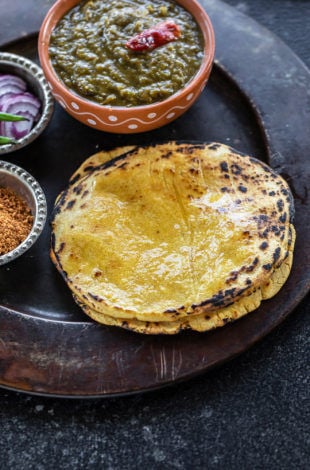 stack of makki ki roti on a plate with a bowl of saag and bowl of powdered jaggery and sliced onions on the sides