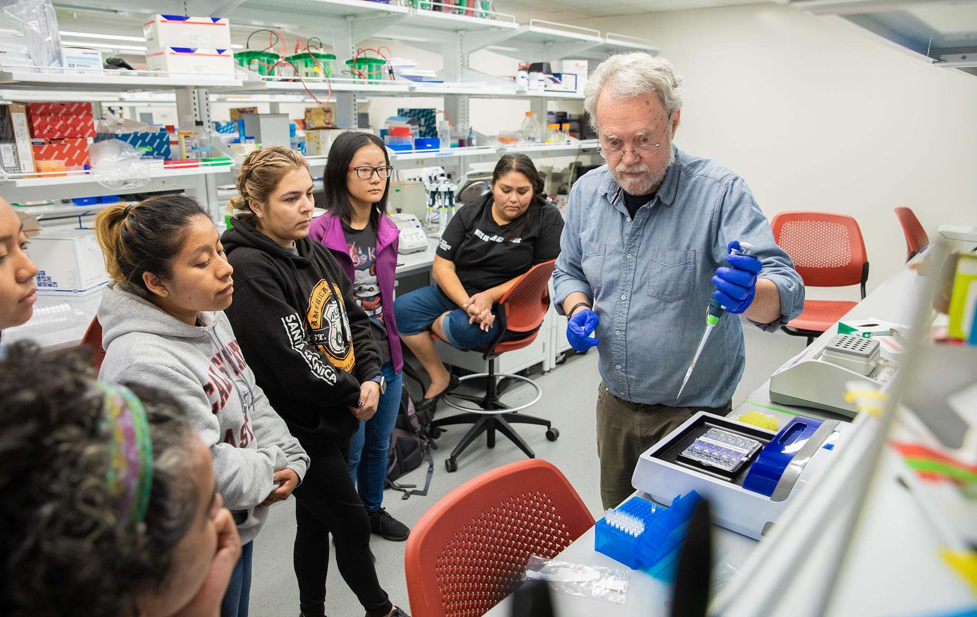 A male professor demonstrates in a science lab while female students watch