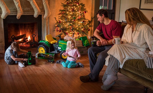 Family on Christmas morning sitting around a fireplace and brightly lit Christmas tree opening John Deere toy tractors and gifts.
