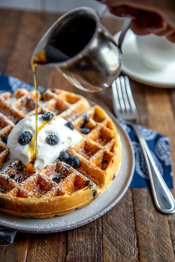 Blueberry waffle with powdered sugar, whipped cream and blueberries with syrup being poured over the top